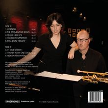 Zwei Menschen am Klavier, einer spielt, die andere lehnt sich über ihn. Instrumente liegen daneben, Umgebung schummrig beleuchtet.
