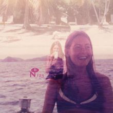 Text: "b N 123". Zwei Frauen, eine lächelt im Vordergrund, die andere trinkt im Hintergrund. Strand und Palmen.