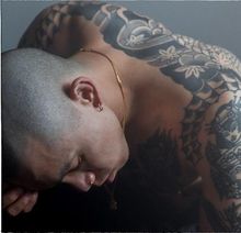 Tätowierter Mensch mit gesenktem Kopf und geschlossenen Augen, trägt eine goldene Halskette.