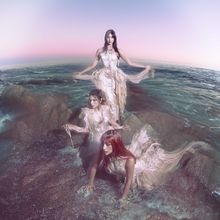 Keine Texte im Bild. Drei Frauen in eleganten, fließenden Kleidern am Meer; mit rosa Himmel im Hintergrund.