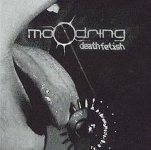 Text: Moodring, Death Fetish. Nahaufnahme eines Mundes, eine Zunge leckt an einem stacheligen Metallring.