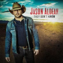 „Jason Aldean, They Don’t Know“. Ein Mann in Jeansjacke und Cowboyhut steht in einer Wüstenlandschaft bei Sonnenuntergang.