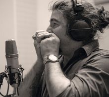 Ein Musiker spielt Mundharmonika, trägt Kopfhörer und steht vor einem Mikrofon. Er trägt eine Uhr und Ringe.