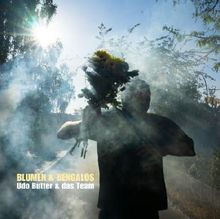 BLUMEN & BENGALOS Udo Butter & das Team. Eine Person trägt Blumen durch Rauch im Sonnenlicht.