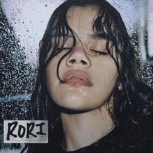 Text "RORI" in stilisiertem Schwarz-Weiß. Gesicht einer Person mit nassem Haar und geschlossenen Augen vor einer regennassen Scheibe.
