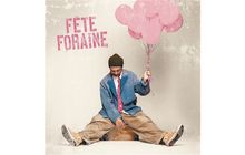 Text: "FÊTE FORAINE". Ein Mann in bunten, lässigen Kleidern sitzt, hält rosa Luftballons und wirkt entspannt und nachdenklich.
