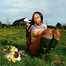 Eine Person sitzt auf Gras mit Blumen daneben, trägt ein buntes Shirt und Stiefel, im Hintergrund ist ein Landhaus.