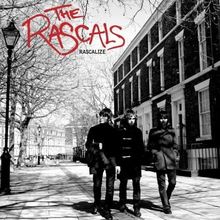"The Rascals RASCALIZE" steht oben. Drei Personen gehen auf einem gepflasterten Gehweg entlang einer Häuserreihe.