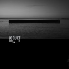 Text: "BE QUIET". Horizontale Struktur im Wasser bei Sonnenuntergang, schwarz-weiße Fotografie.