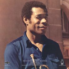 A BYG PRODUCTION. Ein Mann in einem blauen Hemd hält eine Trompete, leicht lächelnd, vor einem unscharfen Hintergrund.