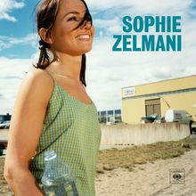 Sophie Zelmani in blauen Buchstaben. Frau in kariertem Kleid mit Wasserflasche. Gelbes Gebäude im Hintergrund, Himmel wolkig.