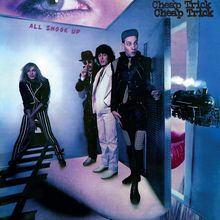 Texte: „ALL SHOOK UP“ und „Cheap Trick“. Vier Männer in auffälliger Kleidung in einem surrealen Raum mit einem Zug und großem Auge.