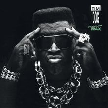 "TIM DOG, PENICILLIN ON WAX." Ein Mann mit Sonnenbrille, ernstem Gesichtsausdruck, auffälligem Schmuck und erhobenen Händen.