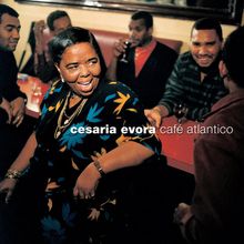 Text: "cesaria evora café atlantico". Eine Frau in bunter Kleidung lächelt in einem lebhaften Café.