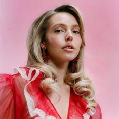 Ich kann keine Texte oder Logos erkennen. Eine Frau in rotem, voluminösem Kleid vor rosa Hintergrund, mit lockigem Haar.