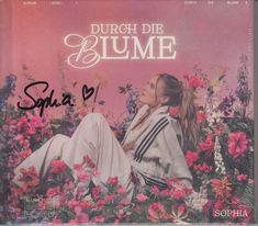 "Durch die Blume," "Sophia" in Schwarz mit Herz. Frau in weißem Outfit sitzt träumerisch in Blumen.