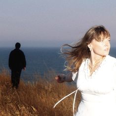 Eine Frau in Weiß steht auf einer Wiese vor dem Meer, der Wind weht in ihr Haar. Ein Mann im Hintergrund.