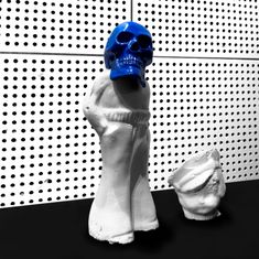Eine abstrakte Skulptur mit einem blauen Totenkopf auf einem weißen Körper, neben einer ähnlichen Form auf schwarzem Boden.