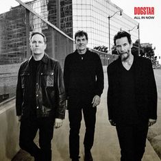 "DOGSTAR ALL IN NOW". Drei Männer in dunkler Kleidung gehen auf einem Gehweg vor urbaner Kulisse.