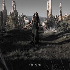 Text: "the dark". Eine Person in schwarzem Kleid steht in einer felsigen Landschaft mit bizarren, hohen Steinformationen.