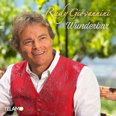 „Rudy Giovannini Wunderbar“ in eleganter Schrift oben. Ein Mann lächelt, trägt eine rote Weste, Hintergrund mit Natur.