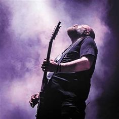 Ein Mann spielt Gitarre in einer dramatischen Pose, umgeben von dichtem, lila beleuchtetem Nebel.
