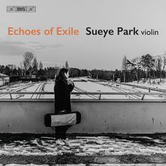 Echoes of Exile, Sueye Park violin. Schwarz-weißes Foto: Frau mit Geigenkoffer steht auf verschneitem Bahnhof.