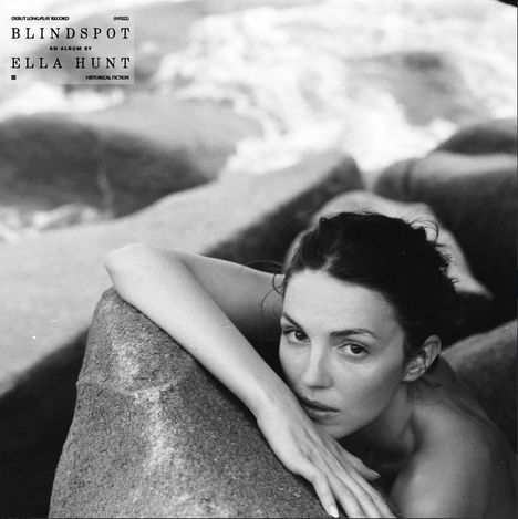 "BLINDSPOT AN ALBUM BY ELLA HUNT" steht links. Eine Frau am Meer lehnt nachdenklich an einem Felsen. Schwarzweißfotografie.