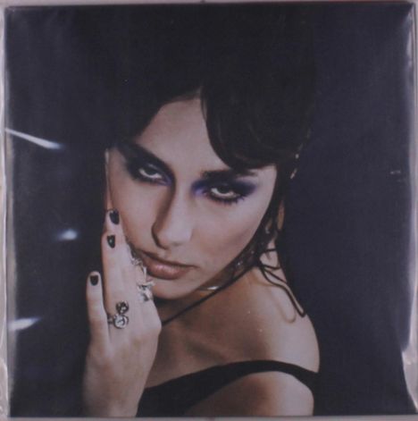 Eine Frau mit dunklem Haar blickt direkt. Sie trägt lila Augen-Makeup, schwarzen Nagellack und auffällige Ringe.