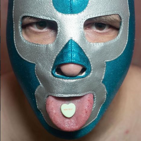 Ein Herz mit der Aufschrift "NEW SELF" auf der Zunge einer Person mit silber-blauer Maske.