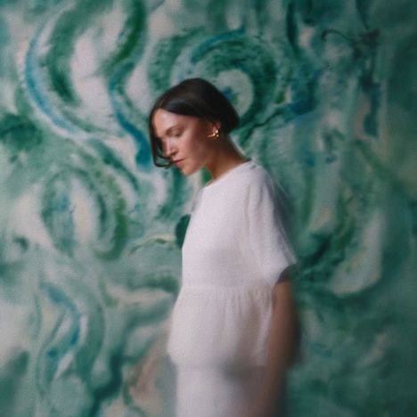 Kein Text erkennbar. Eine Person in Weiß vor einem unscharfen, abstrakten grünen Hintergrund.