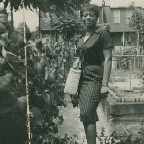 Keine sichtbaren Texte. Eine Frau in einem Garten, trägt ein gestreiftes Kleid und eine Handtasche, lächelt.