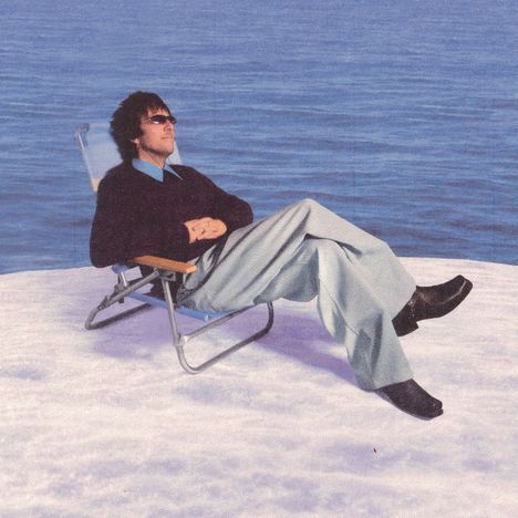 Ein Mann sitzt entspannt in einem Liegestuhl, trägt Sonnenbrille und dunklen Pullover, beobachtet das blaue Meer vor ihm.