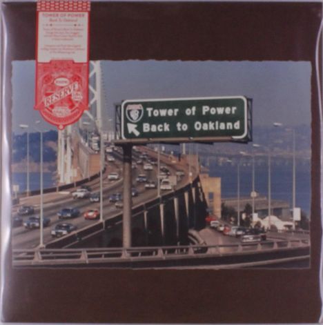 Ein Straßenschild zeigt „Tower of Power Back to Oakland“. Darunter eine befahrene Brücke über Wasser in einer urbanen Umgebung.