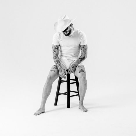Ein Mann in weißer Kleidung und Cowboyhut sitzt auf einem Hocker, voller Tattoos, barfuß, in einem schlichten Raum.