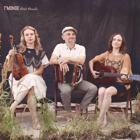 „T'MONDE Petit Paradis“. Drei Personen mit Instrumenten, Geige, Akkordeon, Gitarre, sitzen barfuß auf Stühlen im Freien.