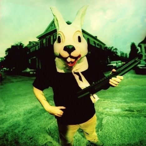 Eine Person in einem Hasenkostüm steht auf der Straße und hält ein Gewehr, im Hintergrund sind Häuser und Bäume.