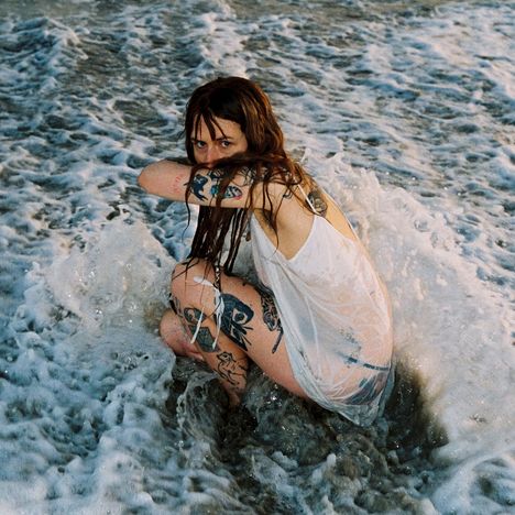 Eine Person mit vielen Tattoos hockt im Wasser, umgeben von Wellen. Sie trägt ein nasses, weißes Oberteil.