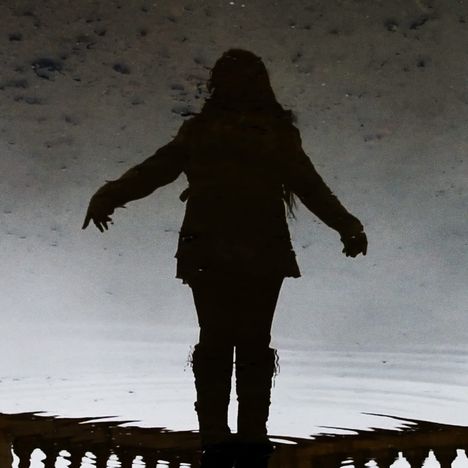 Silhouette einer Person mit ausgestreckten Armen, als Spiegelbild im Wasser, leicht verschwommen.