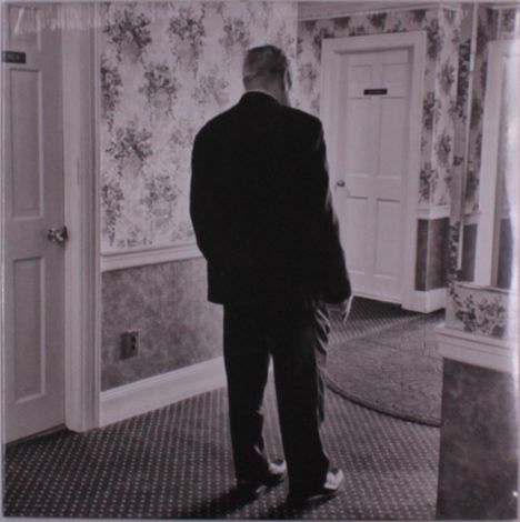 Zwei Türen mit Schildern in einem Flur. Ein älterer Mann im Anzug steht im Raum mit Blumenmuster an den Wänden.