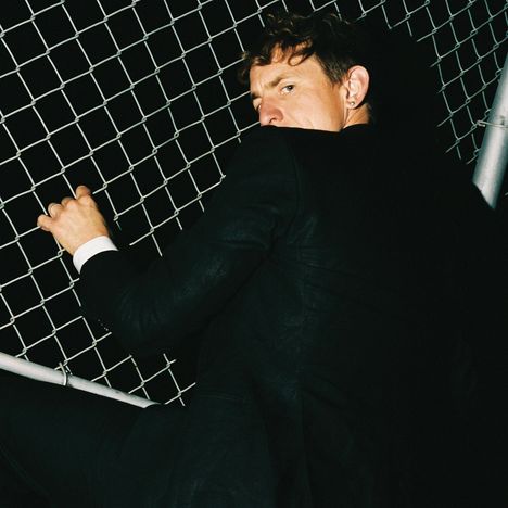 Keine sichtbaren Texte. Eine Person in einem Anzug klettert an einem Maschendrahtzaun in der Dunkelheit.