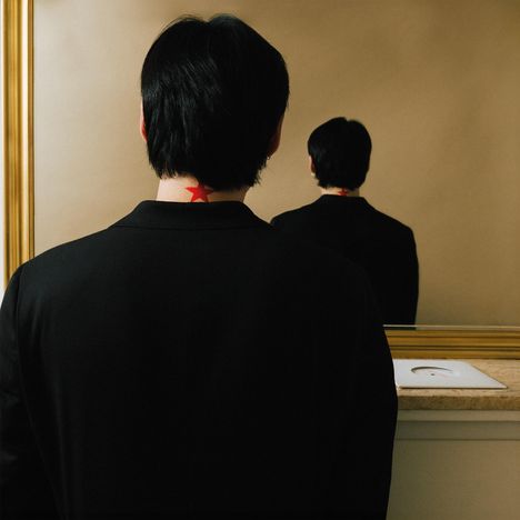 Eine Person mit dunklem Haar und schwarzem Anzug, roter Stern im Nacken, steht vor einem Spiegel.