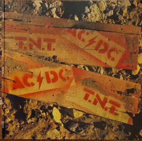 „AC/DC“ und „T.N.T.“ in roter Schrift auf Holzkisten, umgeben von Steinen und Erde.