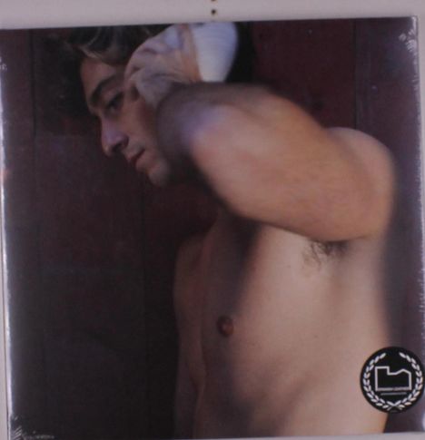 Ein Mann mit Muschel am Ohr, oberkörperfrei, seitlich gezeigt. Unten rechts ist ein schwarzes, rundes Logo mit weißem Symbol.