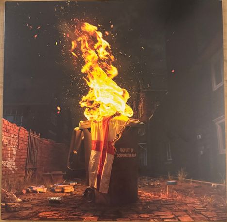 „Property of Corporation Pop“ steht auf einem brennenden Müllcontainer, umgeben von einem dunklen, urbanen Hinterhof.