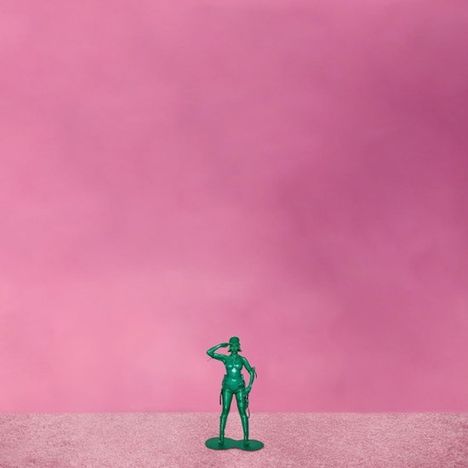 Ein grüner Spielzeugsoldat steht salutierend auf einer rosa Fläche vor einem pinken Hintergrund.