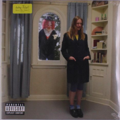 Ein Raum mit einem Fenster, durch das ein Clown lächelt. Eine Frau steht im Raum; Regal mit Büchern und Objekten.