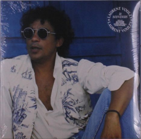 „LAURENT VOULZY 21 SOUVENIRS“ auf blauem Kreis. Person mit Sonnenbrille und gemusterter Weste sitzt lässig.