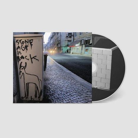 „Stone Age is Back“ steht auf einem Stromkasten. Daneben ist eine Zeichnung eines Tieres. Im Hintergrund eine leere Straße. CD mit Mauer-Motiv.