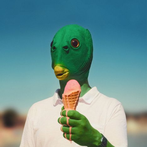 Eine Person in einem grünen Fischkopf-Kostüm hält ein schmelzendes Erdbeereis.